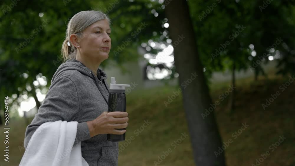 Mature woman drinking water from the sport bottle while walking in the park after training. Elderly female feeling thirsty. Aging youthfully. Mature woman drinking water from the sport bottle while walking in the park after training. Elderly female feeling thirsty. Aging youthfully.