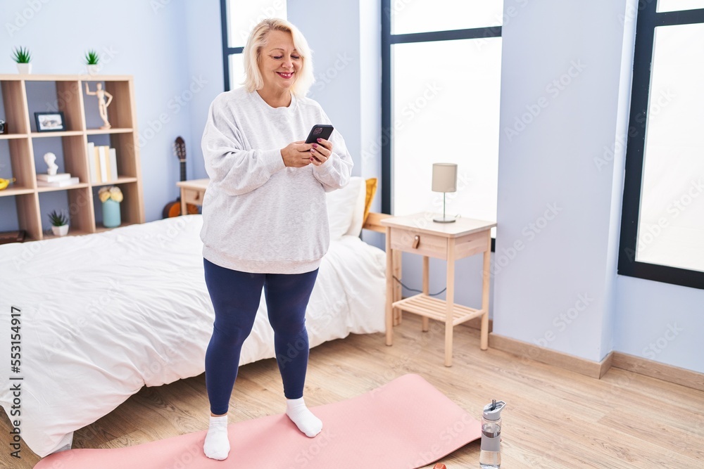 Fototapeta premium Middle age blonde woman using smartphone standing at bedroom