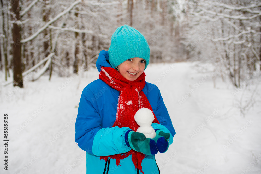 happy boy in winter bright clothes holds a snowball in his hand, walks through the snowy forest