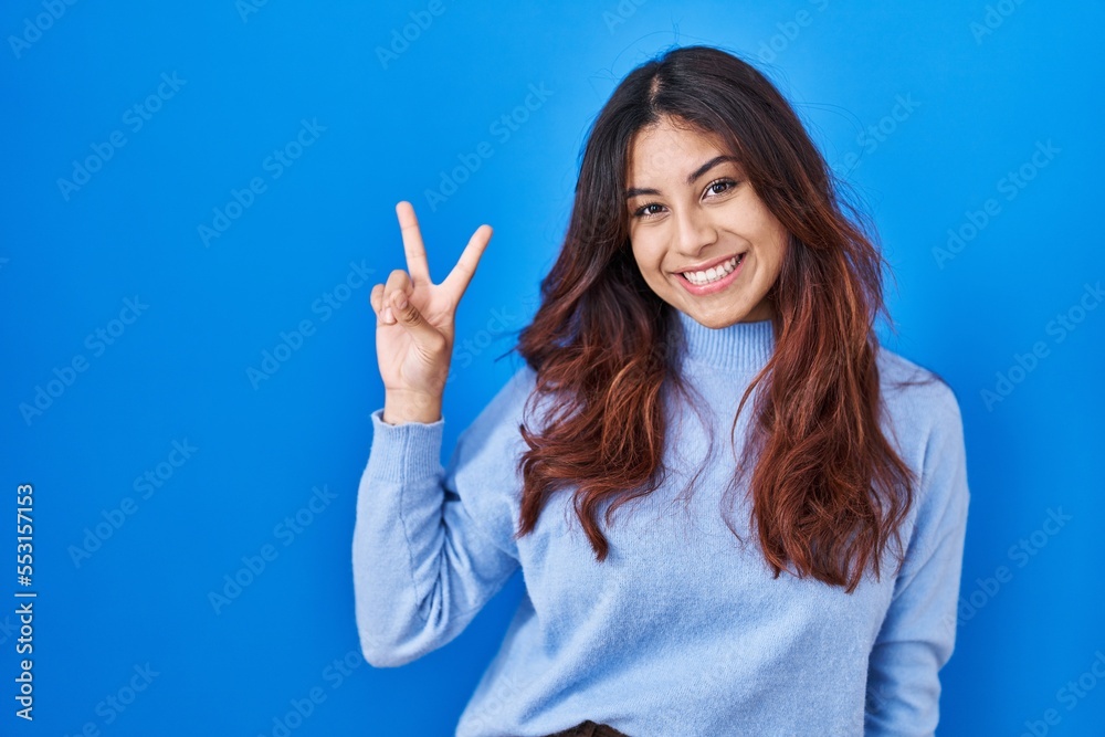Hispanic young woman standing over blue background smiling looking to the camera showing fingers doing victory sign. number two. Hispanic young woman standing over blue background smiling looking to the camera showing fingers doing victory sign. number two.