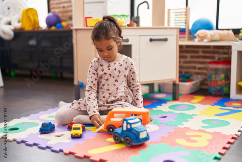 Adorable hispanic girl playing with car toy sitting on floor at kindergarten