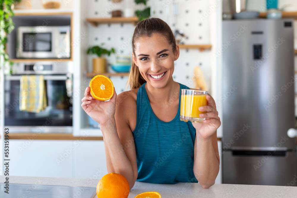 Fototapeta premium Beautiful young woman drinking fresh orange juice in kitchen. Healthy diet. Happy young woman with glass of juice and orange at table in kitchen.