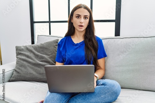 Young brunette woman using laptop at home afraid and shocked with surprise expression, fear and excited face.