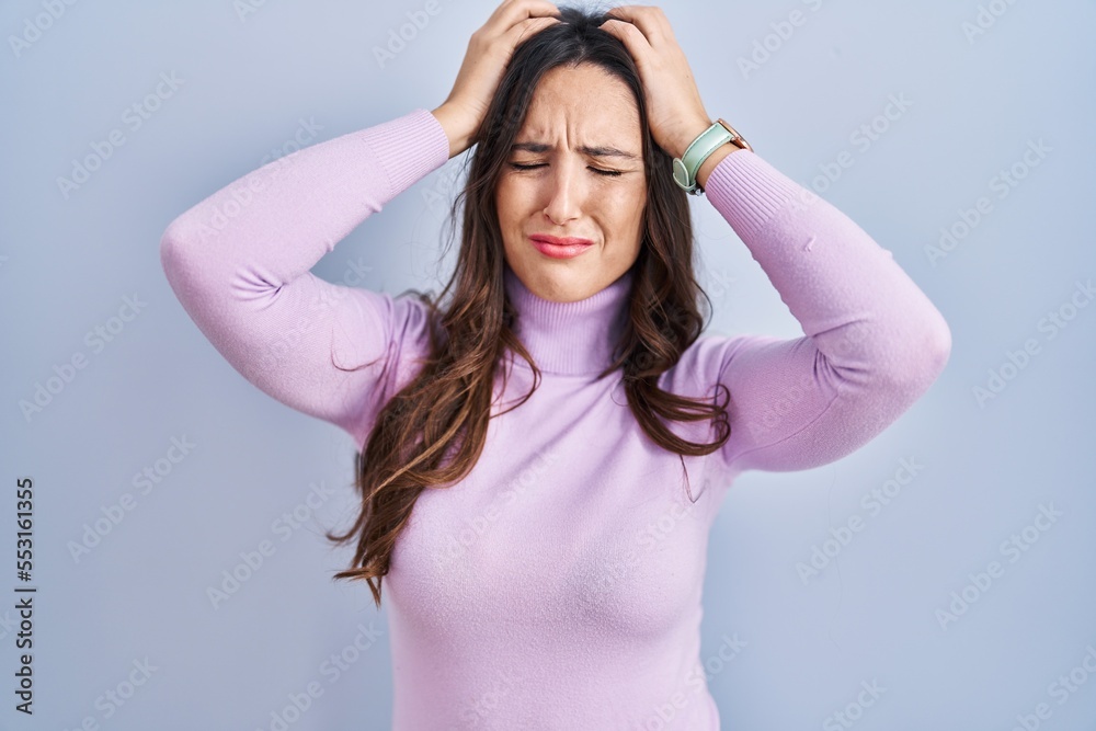 Young brunette woman standing over blue background suffering from headache desperate and stressed because pain and migraine. hands on head. Young brunette woman standing over blue background suffering from headache desperate and stressed because pain and migraine. hands on head.