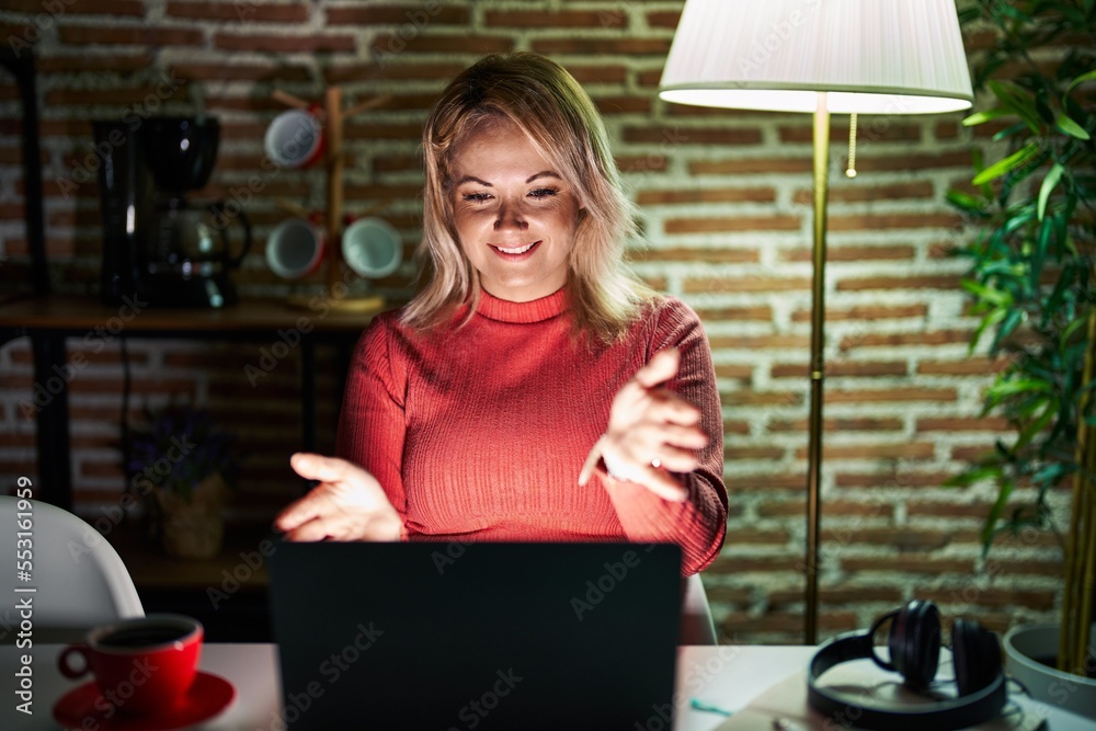 Blonde woman using laptop at night at home looking at the camera smiling with open arms for hug. cheerful expression embracing happiness.