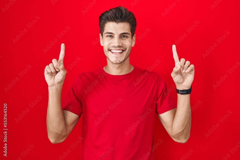 Young hispanic man standing over red background smiling amazed and surprised and pointing up with fingers and raised arms. Young hispanic man standing over red background smiling amazed and surprised and pointing up with fingers and raised arms.