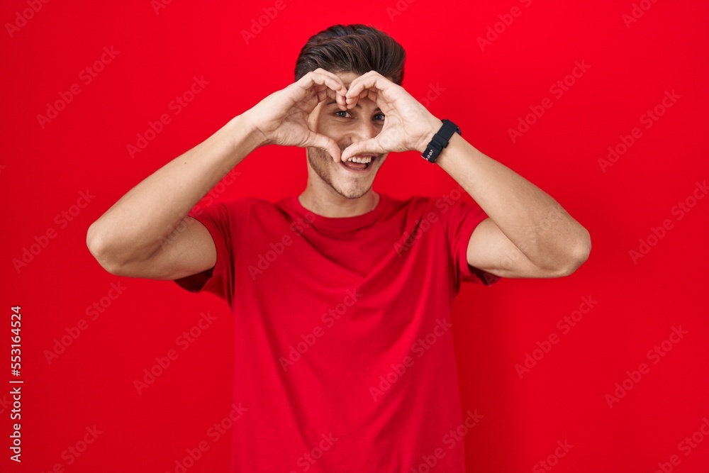 Young hispanic man standing over red background doing heart shape with hand and fingers smiling looking through sign Young hispanic man standing over red background doing heart shape with hand and fingers smiling looking through sign