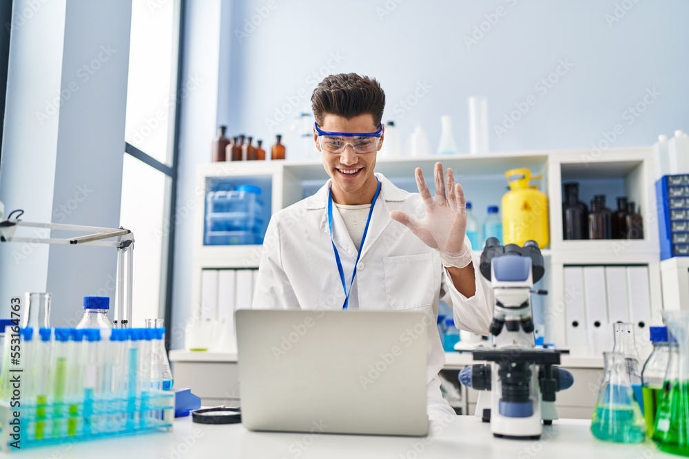 Young hispanic man working at scientist laboratory doing video call looking positive and happy standing and smiling with a confident smile showing teeth Young hispanic man working at scientist laboratory doing video call looking positive and happy standing and smiling with a confident smile showing teeth