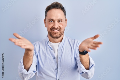 Middle age caucasian man standing over blue background smiling cheerful offering hands giving assistance and acceptance. © Krakenimages.com