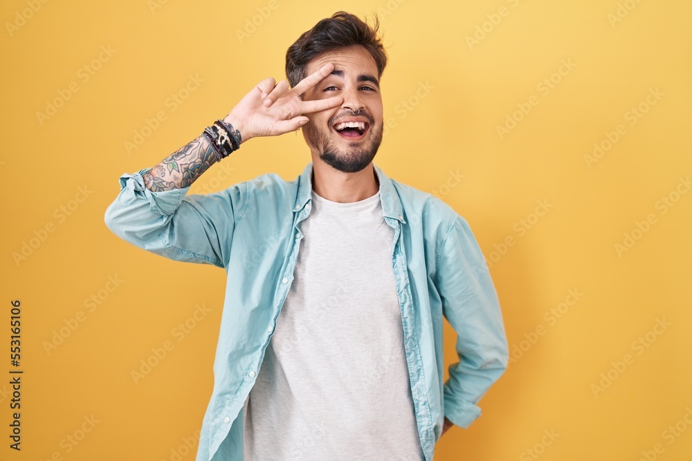 Young hispanic man with tattoos standing over yellow background doing peace symbol with fingers over face, smiling cheerful showing victory Young hispanic man with tattoos standing over yellow background doing peace symbol with fingers over face, smiling cheerful showing victory