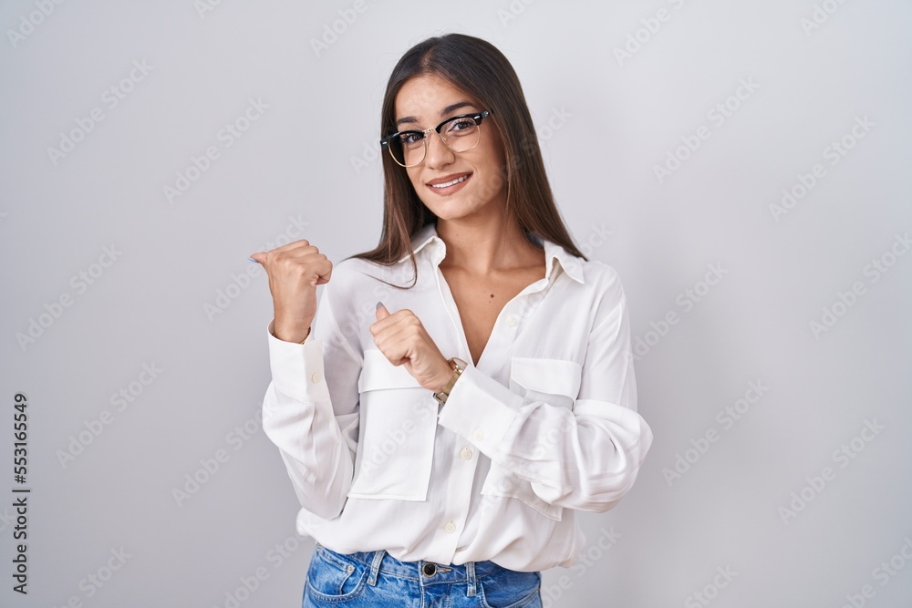 Young brunette woman wearing glasses pointing to the back behind with hand and thumbs up, smiling confident Young brunette woman wearing glasses pointing to the back behind with hand and thumbs up, smiling confident