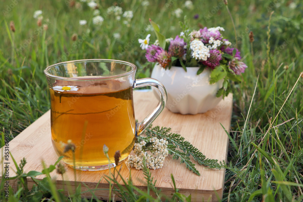 Cup of aromatic herbal tea and ceramic mortar with different wildflowers on green grass outdoors. Space for text