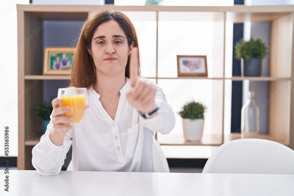 Brunette woman drinking glass of orange juice pointing with finger up and angry expression, showing no gesture Brunette woman drinking glass of orange juice pointing with finger up and angry expression, showing no gesture
