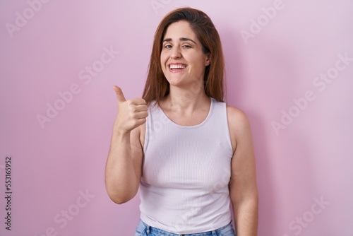 Brunette woman standing over pink background doing happy thumbs up gesture with hand. approving expression looking at the camera showing success.