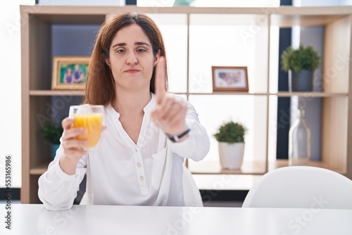 Brunette woman drinking glass of orange juice pointing with finger up and angry expression, showing no gesture