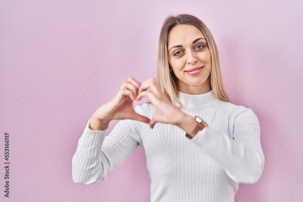 Young blonde woman wearing white sweater over pink background smiling in love doing heart symbol shape with hands. romantic concept. Young blonde woman wearing white sweater over pink background smiling in love doing heart symbol shape with hands. romantic concept.