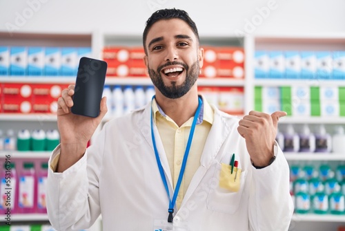Hispanic young man working at pharmacy drugstore showing smartphone screen pointing thumb up to the side smiling happy with open mouth