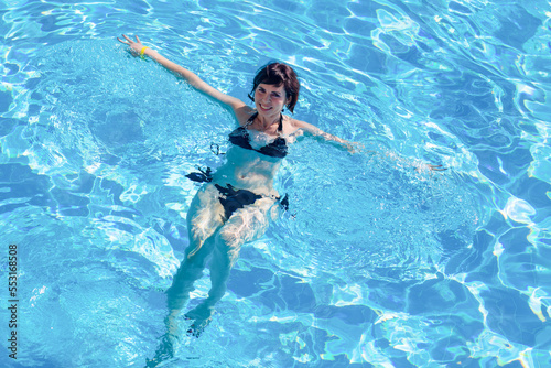 An adult brunette woman is bathing in a pool with blue clear water.