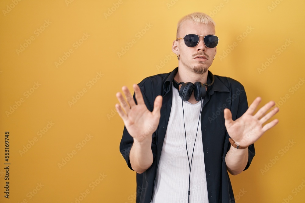 Young caucasian man wearing sunglasses standing over yellow background moving away hands palms showing refusal and denial with afraid and disgusting expression. stop and forbidden.