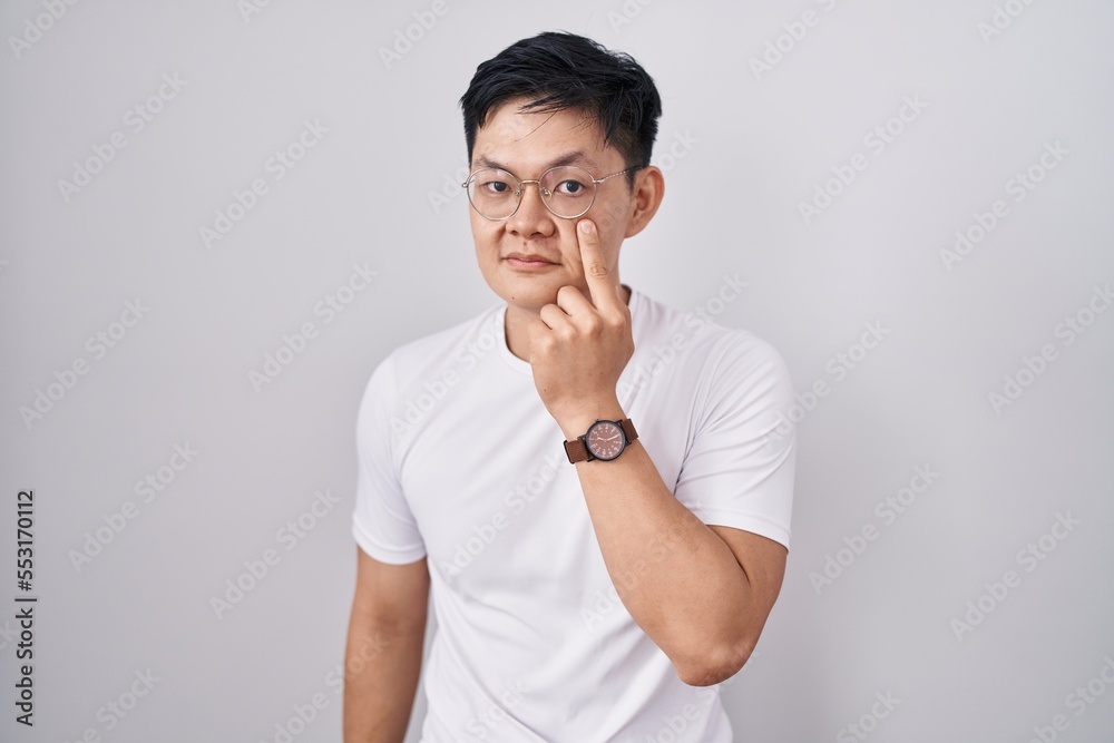 Young asian man standing over white background pointing to the eye watching you gesture, suspicious expression Young asian man standing over white background pointing to the eye watching you gesture, suspicious expression
