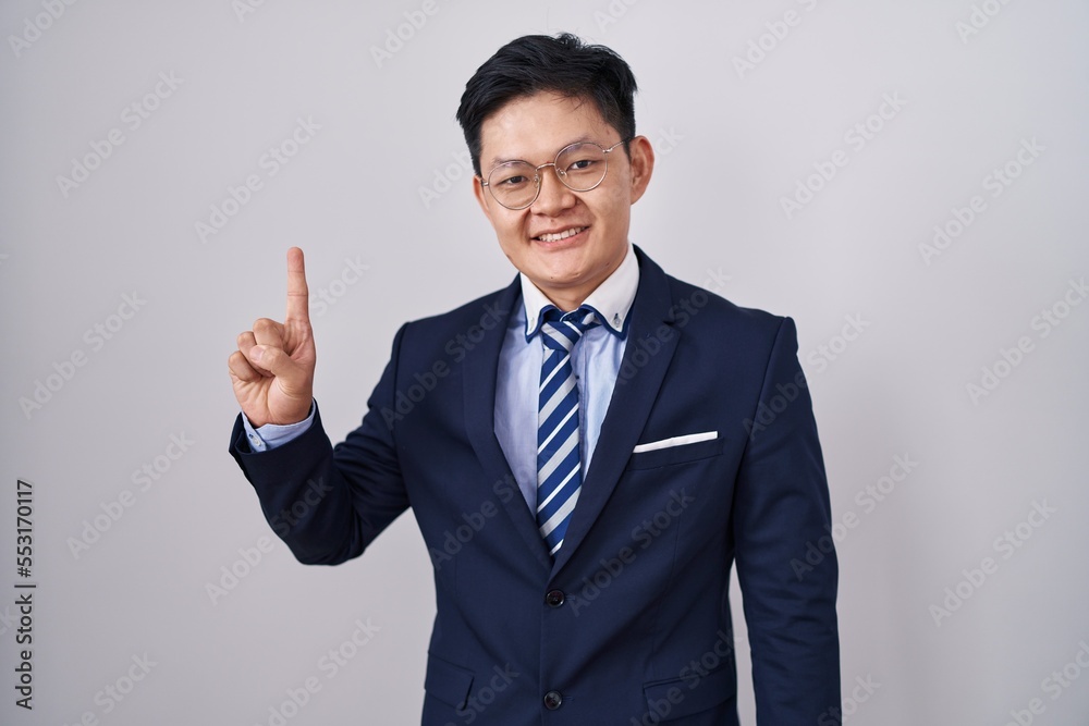 Young asian man wearing business suit and tie showing and pointing up with finger number one while smiling confident and happy. Young asian man wearing business suit and tie showing and pointing up with finger number one while smiling confident and happy.