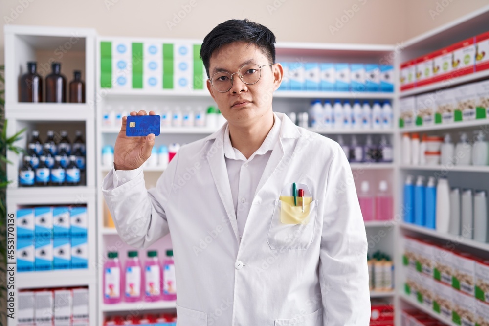Young asian man working at pharmacy drugstore holding credit card thinking attitude and sober expression looking self confident