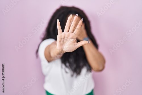 Plus size hispanic woman standing over pink background covering eyes with hands and doing stop gesture with sad and fear expression. embarrassed and negative concept.