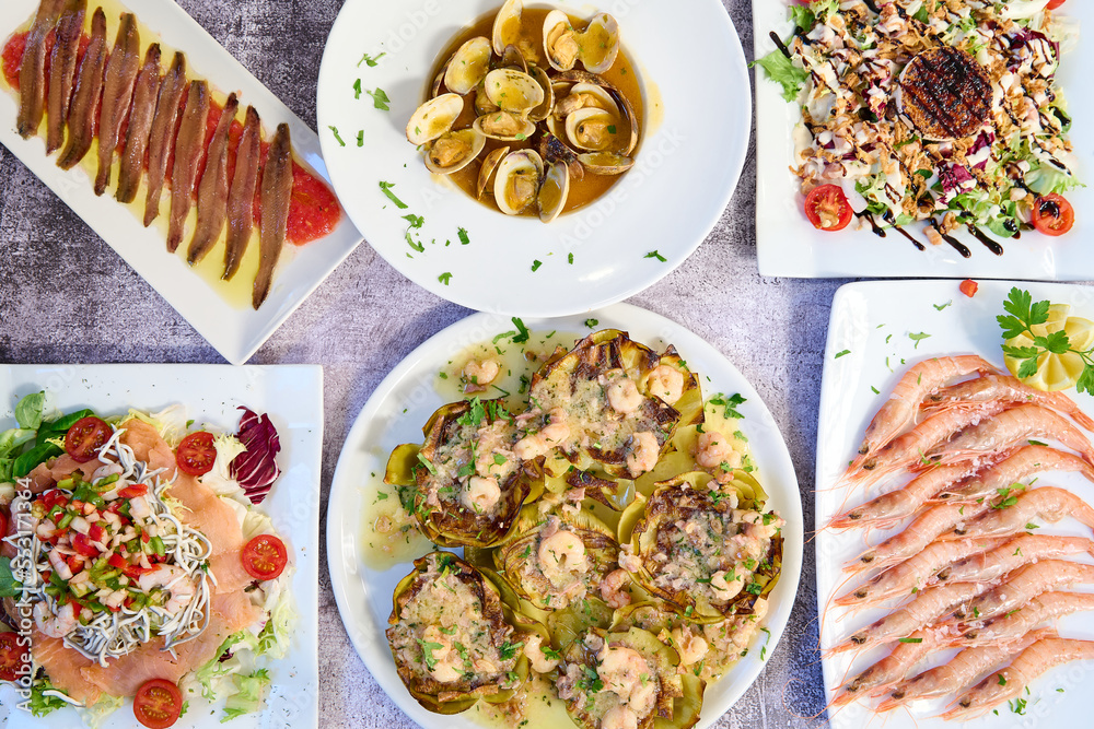 White marble table with typical Spanish food. Grilled white prawns, Cantabrian anchovies, clams, eel salad and artichokes with ham White marble table with typical Spanish food. Grilled white prawns, Cantabrian anchovies, clams, eel salad and artichokes with ham