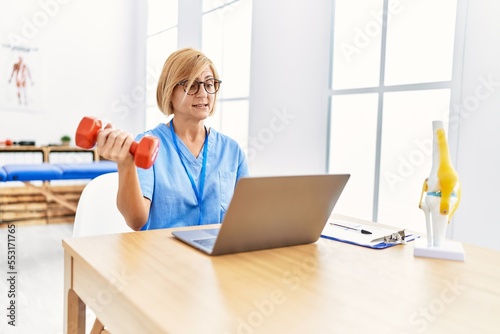 Middle age blonde woman wearing physio therapy uniform doing tele rehab using dumbbells at clinic photo