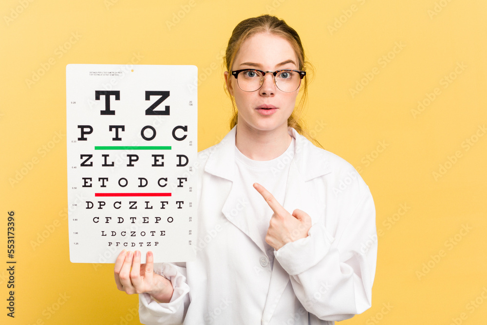 Young caucasian oculist woman holding a eye chart paper isolated on yellow background pointing to the side Young caucasian oculist woman holding a eye chart paper isolated on yellow background pointing to the side