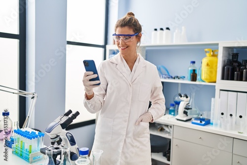 Young blonde woman wearing scientist uniform talking on the smartphone at laboratory