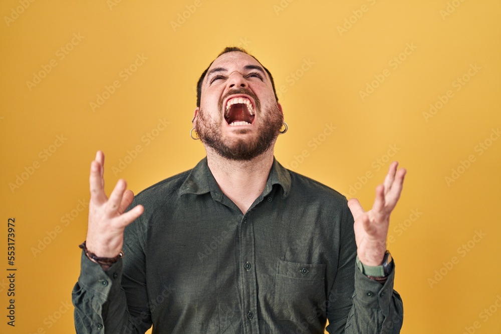 Plus size hispanic man with beard standing over yellow background crazy and mad shouting and yelling with aggressive expression and arms raised. frustration concept. Plus size hispanic man with beard standing over yellow background crazy and mad shouting and yelling with aggressive expression and arms raised. frustration concept.