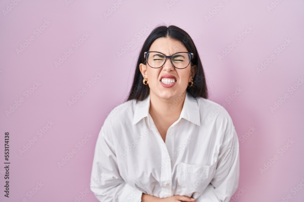 Young brunette woman standing over pink background with hand on stomach because nausea, painful disease feeling unwell. ache concept. Young brunette woman standing over pink background with hand on stomach because nausea, painful disease feeling unwell. ache concept.