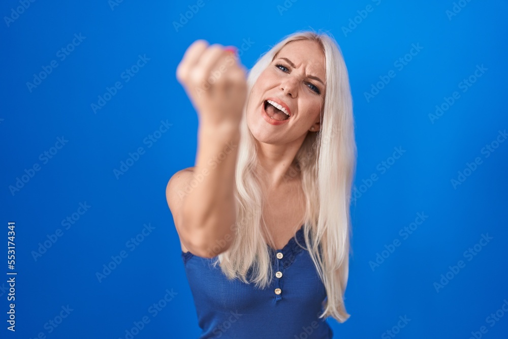 Caucasian woman standing over blue background angry and mad raising fist frustrated and furious while shouting with anger. rage and aggressive concept.