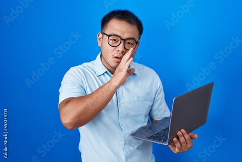 Chinese young man using computer laptop hand on mouth telling secret rumor, whispering malicious talk conversation