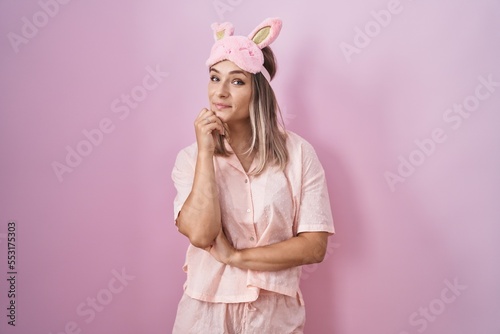 Blonde caucasian woman wearing sleep mask and pajama looking confident at the camera smiling with crossed arms and hand raised on chin. thinking positive.