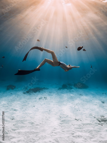 Freediver girl with fins glides underwater in blue transparent ocean with sunset or sunrise rays