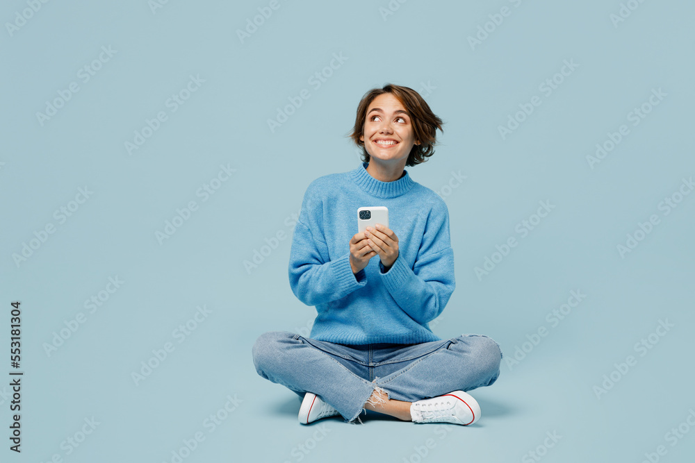 Full body fun young woman wear knitted sweater hold in hand use mobile cell phone look aside on workspace isolated on plain pastel light blue cyan background studio portrait. People lifestyle concept. - obrazy, fototapety, plakaty 