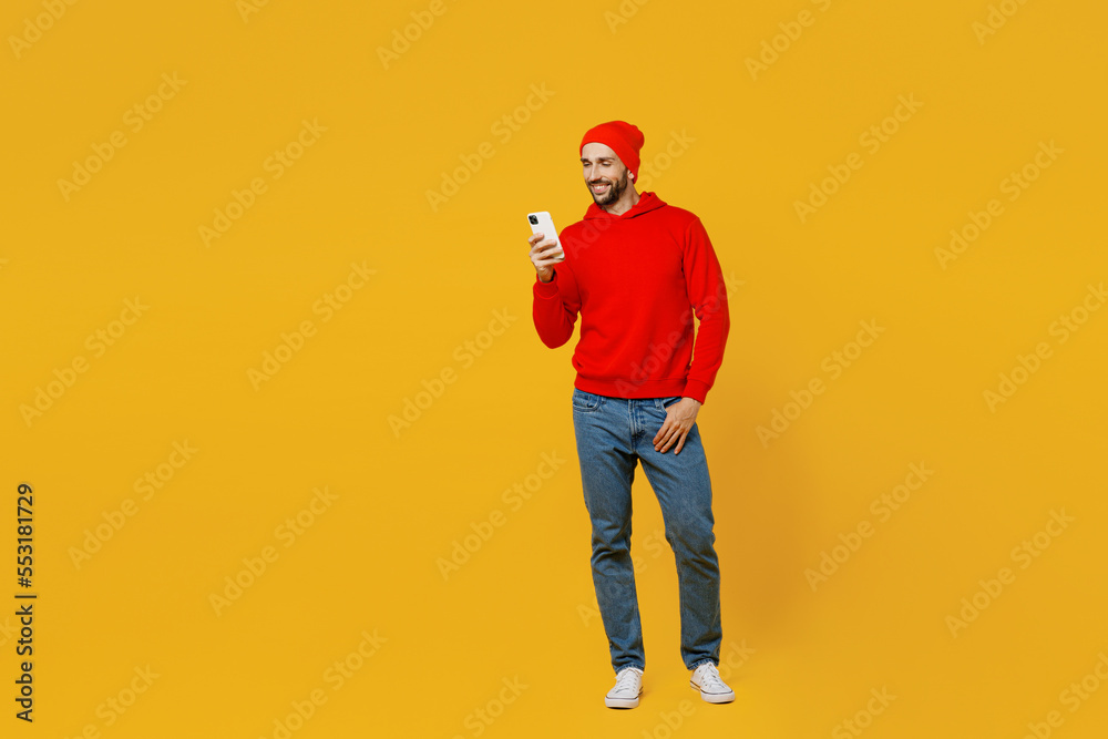 Full body happy smiling fun young caucasian man wear red hoody hat look camera hold in hand use mobile cell phone isolated on plain yellow color background studio portrait. People lifestyle concept. Full body happy smiling fun young caucasian man wear red hoody hat look camera hold in hand use mobile cell phone isolated on plain yellow color background studio portrait. People lifestyle concept.