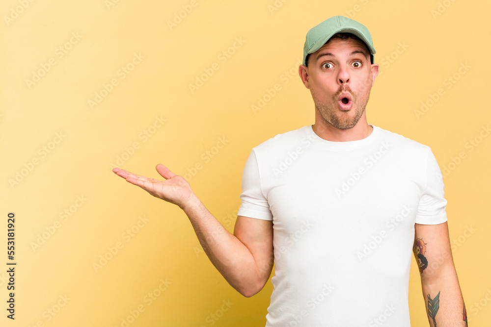 Young caucasian handsome man isolated on yellow background impressed holding copy space on palm.