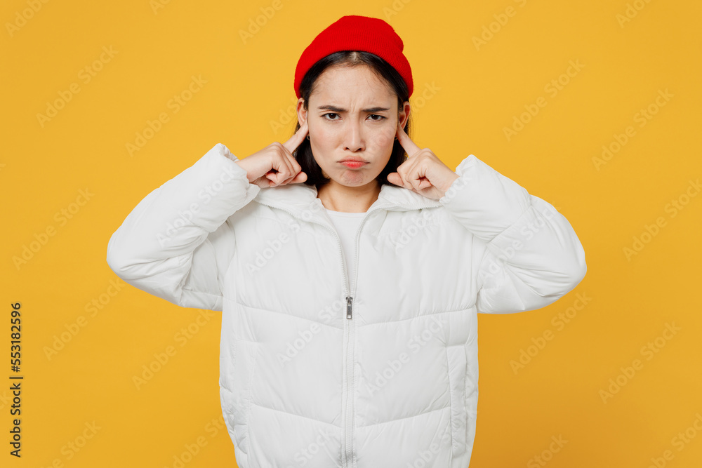 Young stressed sad woman of Asian ethnicity wear white padded windbreaker jacket red hat cover ears with hands fingers do not want to listen scream isolated on plain yellow background studio portrait. Young stressed sad woman of Asian ethnicity wear white padded windbreaker jacket red hat cover ears with hands fingers do not want to listen scream isolated on plain yellow background studio portrait.