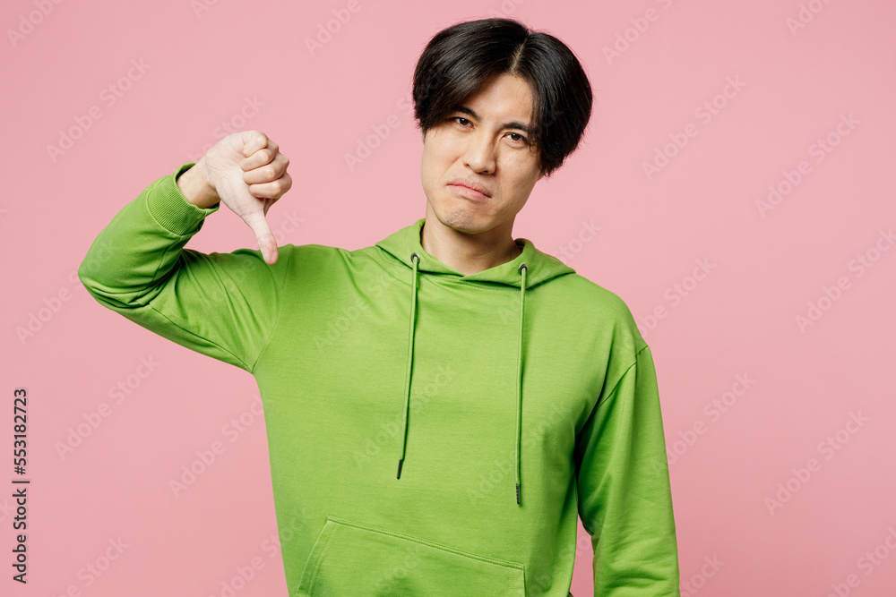 Young unhappy sad man of Asian ethnicity wearing green hoody looking camera showing thumb down dislike gesture isolated on plain pastel light pink background studio portrait. People lifestyle concept. Young unhappy sad man of Asian ethnicity wearing green hoody looking camera showing thumb down dislike gesture isolated on plain pastel light pink background studio portrait. People lifestyle concept.