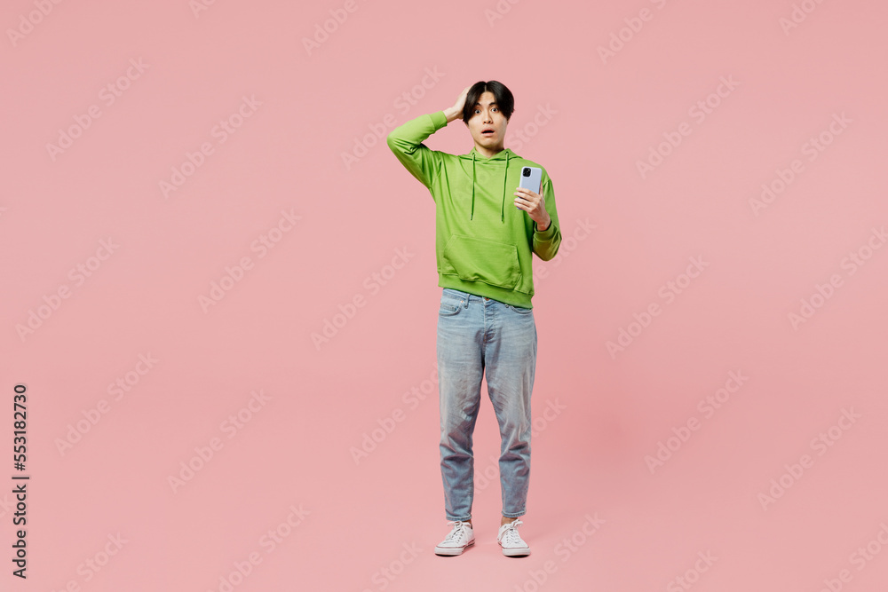 Fototapeta premium Full body shocked sad young man of Asian ethnicity wear green hoody look camera use mobile cell phone hold head isolated on plain pastel light pink background studio portrait People lifestyle concept