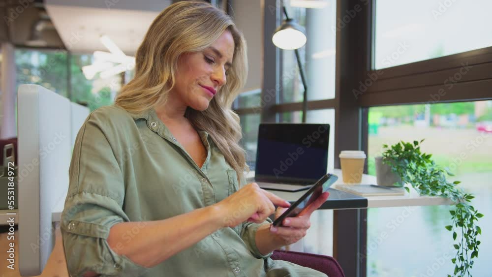 Mature businesswoman taking a break sitting at desk in modern open plan office messaging or browsing on mobile phone and looking out of window - shot in slow motion Mature businesswoman taking a break sitting at desk in modern open plan office messaging or browsing on mobile phone and looking out of window - shot in slow motion
