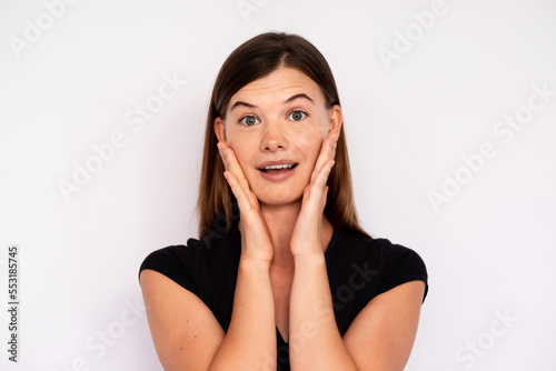 Portrait of excited woman looking at camera with open mouth over white background. Young Caucasian lady wearing black T-shirt and jeans touching face in surprise. Astonishment concept