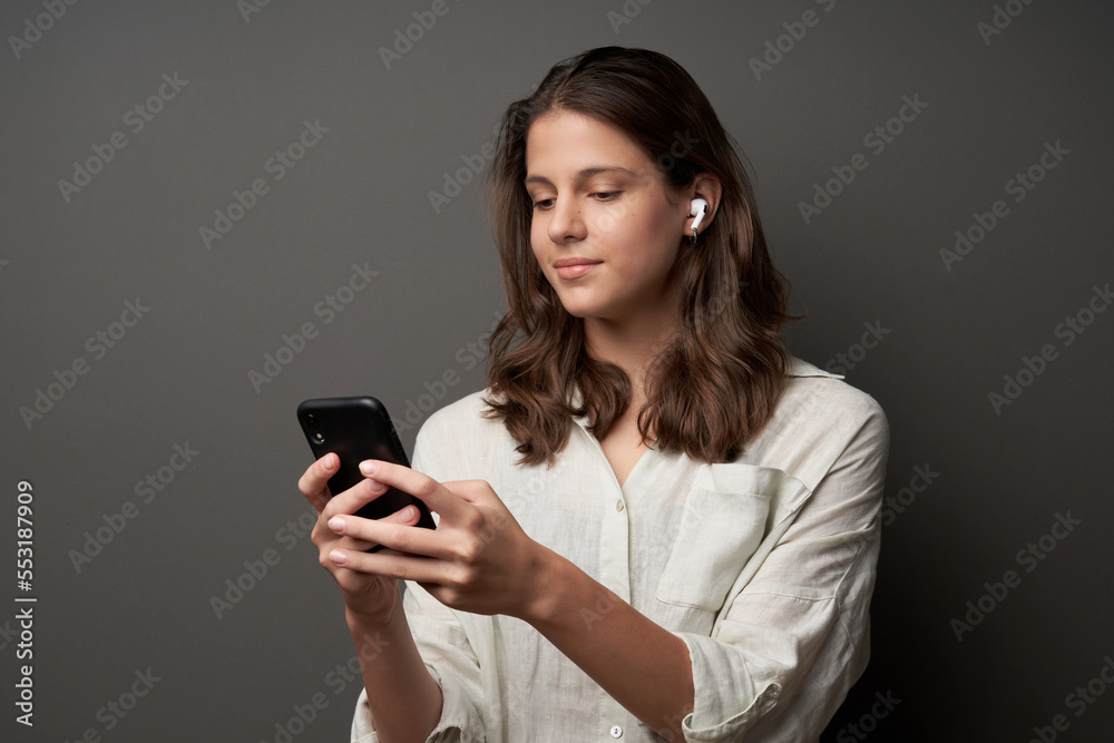 Lovely casual teen girl in earbuds and smartphone in hands, standing on dark background Lovely casual teen girl in earbuds and smartphone in hands, standing on dark background