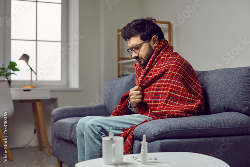 Sick young man in a warm blanket sitting on the sofa at home, feeling cold, freezing, shivering, drinking hot tea and taking medicine. Concept of seasonal cold and flu infection