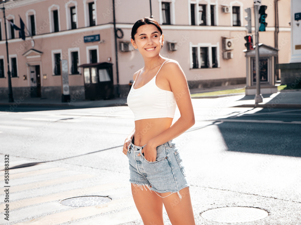 Young beautiful smiling hipster woman in trendy summer white top and shorts clothes. Sexy carefree model posing in the street at sunny day. Positive brunette female. Cheerful and happy Young beautiful smiling hipster woman in trendy summer white top and shorts clothes. Sexy carefree model posing in the street at sunny day. Positive brunette female. Cheerful and happy
