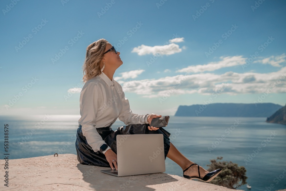 Freelance women sea. She is working on the computer. Good looking middle aged woman typing on a laptop keyboard outdoors with a beautiful sea view. The concept of remote work.