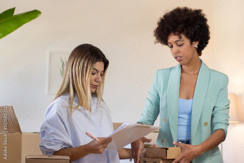 Obraz premium Postal coworkers examining numbers of parcels. Caucasian woman holding pen and clipboard while blackwoman stacking boxes. Logistics concept